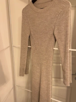 Beige ribbad långklänning - Beige ribbad långklänning med lång ärm och rund hals. Klänningen har en kroppsnära passform och är tillverkad i ett mjukt stickat material som ger en bekväm känsla. Perfekt för dig som gillar stilrena och enkla plagg. Helt oanvänd, passar både Xs och S