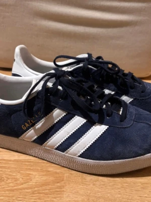 Adidas Gazelle mörkblå sneakers - Säljer ett par Adidas Gazelle i marinblått med vita ränder. Använda bara ett fåtal gånger och i riktigt bra skick. Inga fula märken eller slitningar, ser nästan nya ut (se bilder). Pris går att diskutera, samt hör av om fler bilder önskas😊