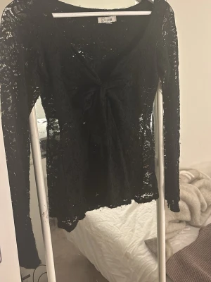 Svart spetsblus med knut från Brisli - Snygg svart långärmad topp i spets från Brisli. Modellen har djup urringning och en knut framtill. Hela toppen är genombruten med blommigt spetsmönster och har en feminin, figurnära passform. Perfekt för att lyfta vilken outfit som helst. (Har defekter på toppen)