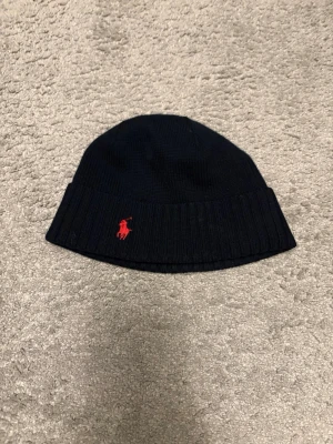 Marinblå mössa från Polo Ralph Lauren - Snygg marinblå ribbstickad mössa från Polo Ralph Lauren med klassisk röd logga broderad framtill. Mössan har uppvikt kant och är tillverkad i mjukt stickat material som håller dig varm under kalla dagar.