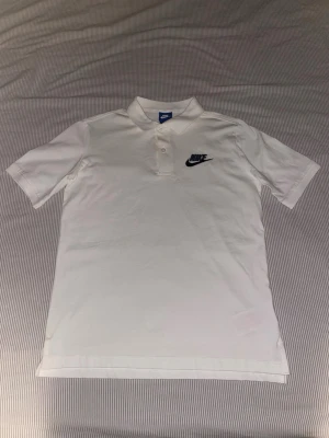 Vit pikétröja från Nike - Klassisk vit pikétröja från Nike med broderad logga på bröstet. Tröjan har korta ärmar, krage och knappar framtill. Tillverkad i mjuk bomull som känns skön mot huden. Perfekt för en clean och sportig look.