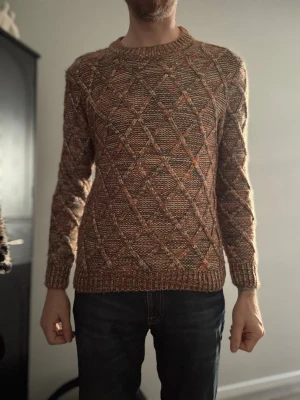 Brunmelerad stickad tröja med mönster - Säljer en brunmelerad stickad tröja med rutmönster i beige, brunt och inslag av orange. Tröjan har rund halsringning, långa ärmar och ribbstickade muddar vid ärmslut och nederkant. Perfekt för dig som gillar retrovibbar och unika detaljer.