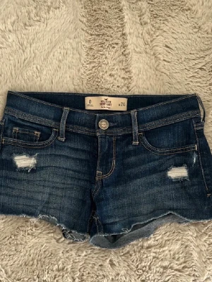 Mörkblå jeansshorts från Hollister - Snygga mörkblå jeansshorts från Hollister med slitna detaljer och rå kant nertill. Klassisk femficksmodell med kontrastsömmar och låg midja. Perfekta för varma dagar och en avslappnad look.