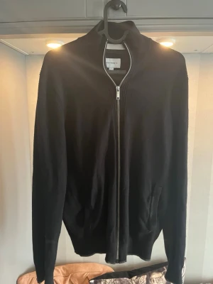 Svart ziptröja  - Klassisk svart ziptröja  med hög krage och hel dragkedja framtill. Tröjan har långa ärmar och ribbade muddar vid ärmslut och nederkant. Perfekt att slänga på sig över en t-shirt för en clean look.