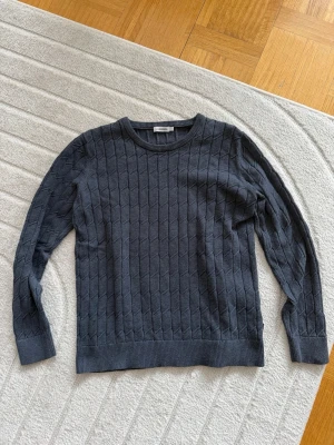 Mörkgrå kabelstickad tröja från J.Lindberg - Snygg mörkgrå kabelstickad tröja från J.Lindberg. Den har rund halsringning och långa ärmar. Tröjan har ett diskret mönster och ribbade muddar vid ärmslut och nederkant. Perfekt för lager på lager och enkel att matcha med olika outfits. Tröjan är en L men passar som en M