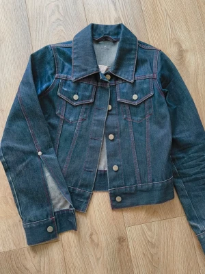 Vintage mörkblå jeansjacka från Action Jeans (Acne) - Snygg mörkblå jeansjacka från Action Jeans med röda kontrastsömmar och klassisk krage. Jackan har två bröstfickor med lock och silverfärgade knappar framtill. Perfekt för dig som vill ha en tidlös och cool look.
