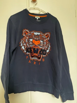 Mörkblå Kenzo sweatshirt med tiger - Säljer en mörkblå sweatshirt från Kenzo. Tröjan har rund halsringning och ribbade muddar. Materialet är mjuk bomull som känns skön mot huden. Perfekt för dig som vill sticka ut med en fet design.