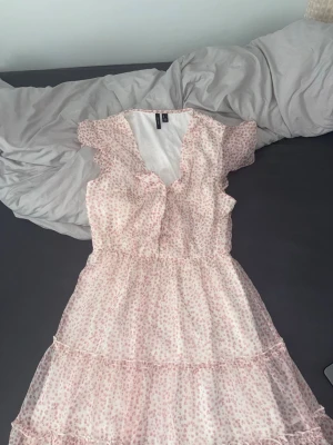 Blommig volangklänning Vero Moda - Söt kortklänning från Vero Moda i ljusrosa och vitt med blommigt mönster. Klänningen har volangdetaljer, v-ringning och korta ärmar. Tillverkad i ett lätt och luftigt material, perfekt för varma dagar.