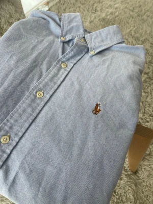 Polo Ralph Lauren Skjorta Ljusblå - En riktigt schysst skjorta från Ralph Lauren i en härlig, ljusblå färg.  Skick: Utmärkt, inga skador eller skavanker såsom hål eller fläckar.  Mått: • Axel - Axel: 44cm • Axel - Midja: 61 cm • Armhåla - Armhåla: 49cm  Detaljer • Tillverkad i 100% Bomull • Justerbara ärmar • Broderad logga på vänster sida av bröstet • Ljusblå färg, fint till sommaren.  Nypris: 1995kr - Mitt Pris: 449kr Skickas inom 24 timmar efter köp!