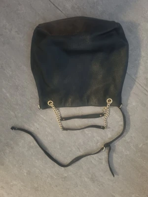 Svart väska med kedjedetaljer från Michael Kors - Stilren svart väska med mjukt konstläder med guldfärgade kedjor och öglor. Väskan har justerbar axelrem i läderimitation och en rymlig form. Perfekt för dig som gillar en enkel men trendig look. Axelremmen behöver lagas därav priset. Väskan är äkta