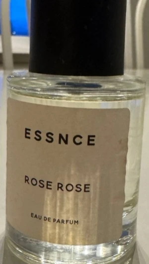 Essnce Rose Rose Eau de Parfum - En elegant parfym från Essnce med doften Rose Rose. Flaskan är rund och genomskinlig med en enkel beige etikett och svart kork. Innehållet är klart och flaskan har en stilren, minimalistisk design som passar perfekt på parfymhyllan.