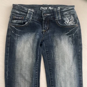 Blå 2000s tal lågmidjade jeans från crazy age - Snygga blå jeans från Crazy Age med slitningar och broderade vita fåglar på både fram- och bakfickor. 