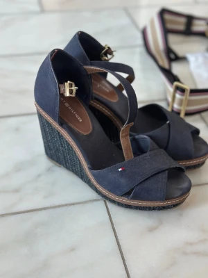 Marinblå kilklackssandaler Tommy Hilfiger - Snygga marinblå sandaler med kilklack från Tommy Hilfiger. Skorna har korsade remmar framtill, justerbart spänne i guld och en detalj med logga på remmen. Ovandelen är i textil och sulan har flätad look. Perfekta för en trendig och bekväm stil.