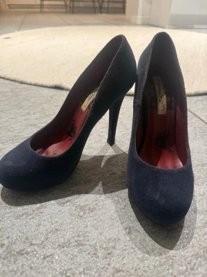 Svarta pumps från Ed Hardy - Snygga svarta pumps från Ed Hardy med rundad tå och hög klack. Utsidan är i mockaliknande material och insidan har ett rött foder med tryckt mönster och logga. Perfekta för dig som vill ha en klassisk men ändå edgy look.