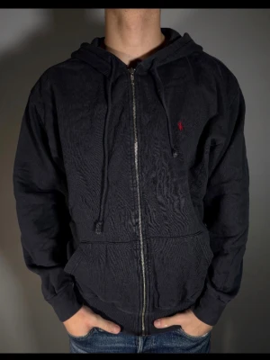 Svart zip hoodie från Ralph Lauren - Säljer en svart hoodie med dragkedja från Ralph Lauren. Klassisk design med huva och dragsnören, samt fickor framtill. Liten röd Polo-logga broderad på bröstet. Tillverkad i mjukt bomullsmaterial som är skönt att bära.