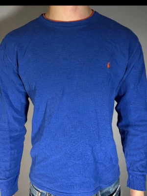 Blå långärmad tröja från Ralph Lauren - Blå långärmad tröja från Ralph Lauren med rund halsringning och orange broderad logga på bröstet. Tröjan har en diskret struktur i materialet och orange detalj vid halsen. Perfekt för en avslappnad och stilren look.