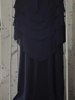 Abaya med khimar  - Säljer en elegant långklänning Jilbab med khimar med flera lager volang upptill och långa ärmar. Finns i färgerna vit, beige, brun, grå, marinblå och svart. Klänningen har en loose passform och är tillverkad i ett lätt syntetmaterial som faller snyggt. 