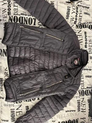 Mörkgrå pufferjacka med dragkedjor - mörkgrå pufferjacka med flera dragkedjeförsedda fickor och hög krage med spänne. Jackan har quiltad design och detaljer i svart samt metallknappar på kragen. Den är sliten runt dragkedjan till fickorna 