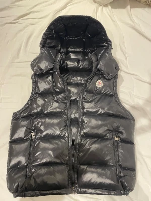 Svart dunväst från Moncler med huva - Snygg svart dunväst från Moncler med glansig finish och stor vadderad huva. Västen har två dragkedjeförsedda öfickor fram och klassisk Moncler-logga på bröstet. Perfekt för lager på lager och riktigt street. Material: dunfyllning och nylon.