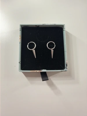 Silvriga örhängen med spetsig detalj - Säljer ett par silvriga örhängen i form av små ringar med hängande spetsiga koner. Örhängena har en minimalistisk och edgy design som sticker ut. Perfekta för dig som gillar en stilren men ändå tuff accessoar.