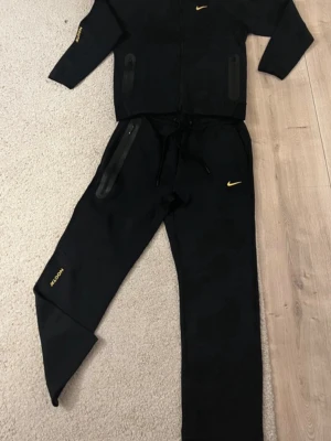 Svarta NOCTA x Nike mjukisbyxor - Säljer ett par svarta mjukisbyxor från Nike och NOCTA-kollektionen. Byxorna har gul NOCTA-logga på benet och en liten gul Nike Swoosh vid fickan. Utrustade med dragkedjefickor och snörning i midjan. Perfekta för en chill och sportig look.