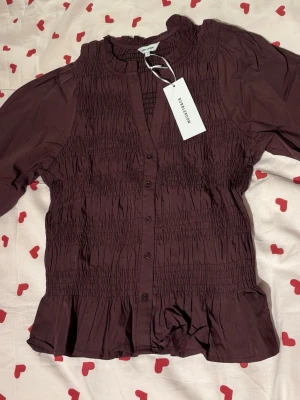 Plommonlila blus från Bubbleroom - Trendig vinröd blus från Bubbleroom med smockdetaljer över hela kroppen, knappar framtill och volangkant nedtill. Blusen har puffiga långärmar och en rundad halsringning. Storlek S, aldrig använd 🥰