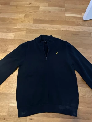 Svart halvzip tröja från Lyle & Scott - Snygg svart långärmad tröja från Lyle & Scott med halv dragkedja och klassisk gul logga på bröstet. Tröjan har en rak passform och ribbade muddar vid ärmslut och nederkant. Perfekt till en clean och stilren look.