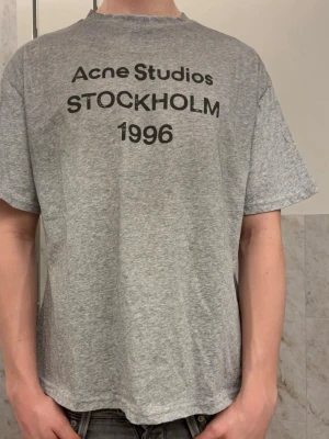 Grå Acne Studios t-shirt - Helt ny t-shirt från Acne Studios med svart text 'Acne Studios STOCKHOLM 1996' på bröstet. T shirten är helt ny och tag finns kvar. Storleken är S men den sitter relaxed så den passar M. 