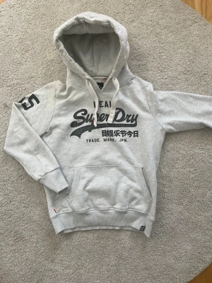 Ljusgrå Superdry hoodie med tryck - Snygg ljusgrå hoodie från Superdry med svart logga och japanska tecken på bröstet. Stor ficka framtill, huva med dragsko och tryckt siffra på ärmen. Perfekt för en avslappnad och trendig stil.
