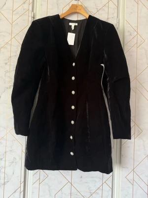 Provkollektion svart sammetsklänning med knappar - Denna är troligtvis från en provkollektion som har hängt ute på en mässa. Stilren svart Sasha sammetsklänning med v-ringning och långa ärmar. Klänningen har dekorativa knappar framtill och en figurnära passform som ger en elegant siluett. Läger ut igen då jag har två likadana klänningar. I material recycled polyester.