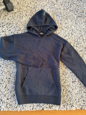 INDICODE Hoodie - Mysig mörkgrå stickad hoodie från INDICODE med stor huva och magficka. Tröjan har ribbade muddar vid ärmslut och nederkant, vilket ger en schysst passform. Perfekt för dig som gillar en enkel och stilren look med lite extra komfort.