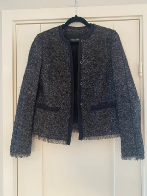 Svart och vit tweedkavaj från Dolce & Gabbana - Svart och vit mönstrad kortare jacka i strl 38 fr Dolce & Gabbana.  Den har en knappstängning framtill och är designad med en rak passform och fransade kanter vid ärmslut och nederkant. Jackan har två framfickor och är fodrad på insidan.  Sparsamt använd och i fint begagnat skick   Mått Över bröstet: ca 45 cm Längd från axeln och ner: ca 56 cm  62% ull, 20% silke, 7% alpacka, 7% mohair och 4% polyamid   Kommer från ett djur och rökfritt hem 