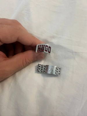 Två silvriga ringar från HUGO - Säljer två stilrena ringar från HUGO. Den ena ringen är bred och har en böjd form med HUGO-loggan i rött och vitt framtill. Den andra ringen har en länkdesign i silverfärg och HUGO-loggan ingraverad. Perfekta accessoarer för dig som gillar statement-smycken.