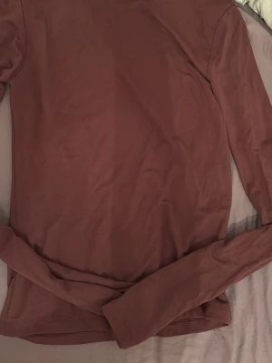 Mörkbrun långärmad topp med öppen rygg - Säljer en mörkbrun långärmad topp med snygg öppen rygg. Toppen har en enkel, tight passform och är perfekt för dig som gillar stilrena plagg med en twist. Materialet känns mjukt och stretchigt, vilket gör den bekväm att bära. Aldrig använd. 