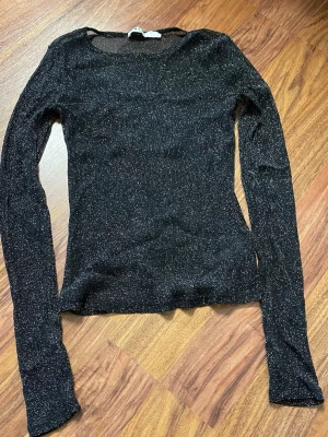 Svart glittrig långärmad topp NA-KD - Snygg svart långärmad topp från NA-KD med glittrigt material som ger en festlig känsla. Tight passform och rund halsringning. Perfekt att styla med jeans eller kjol för en trendig look.