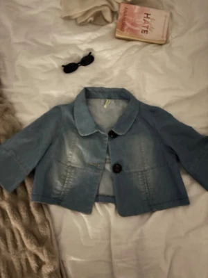 Kort ljusblå jeansjacka med stora knappar - Säljer en trendig kort jeansjacka/jeansbolero i ljusblå denim med stora svarta knappar framtill och bred krage. Jackan har trekvartsärmar som ger en cool retrovibe. Perfekt till våren och sommaren💞