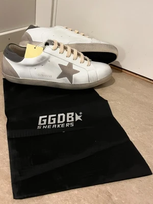 Vita GGDB Superstar sneakers - Snygga vita GGDB Superstar sneakers med beige mockadetaljer och grå stjärna på sidan. Skorna har beige snören, grå häl och klassisk låg siluett. Tillverkade i läder och mocka för en trendig och avslappnad look. Perfekta för dig som gillar streetstyle och exklusiva märken.