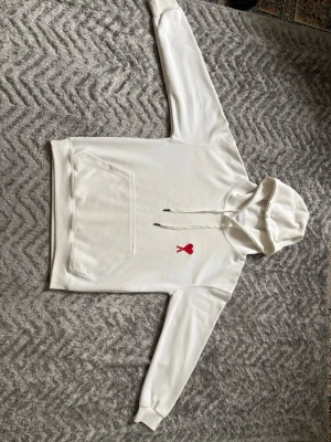 Vit hoodie från Ami Paris med rött hjärta - Säljer en stilren vit hoodie från Ami Paris med det ikoniska röda hjärtat broderat på bröstet. Hoodien har huva med dragsko, magficka och ribbade muddar. Perfekt för dig som gillar enkel men snygg streetwear. Materialet känns mjukt och skönt.