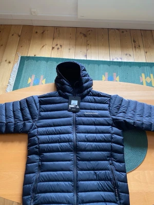 Svart dunjacka med huva Peak Performance - Snygg svart dunjacka från Peak Performance med huva och dragkedja framtill. Jackan har två sidofickor med dragkedja och diskret logga på bröstet. Lättviktig och quiltad design som håller värmen under kalla vinterdagar.