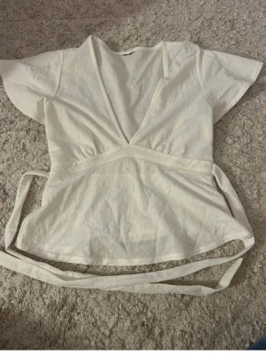 Vit omlottopp med knytband - Säljer en vit omlottopp med korta ärmar och breda knytband i midjan. Toppen har v-ringning och är tillverkad i ett mjukt bomullstyg. Perfekt för en avslappnad och trendig look.