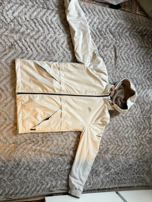 Beige vindjacka med huva - Snygg beige vindjacka från Athletic Sports med dragkedja framtill, två snedställda fickor och justerbar huva med dragsko. Jackan har ribbade muddar vid ärmsluten och tryckt logga på ryggen. Perfekt för blåsiga dagar och enkel att matcha.