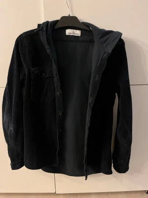 Svart/mörkblå manchester overshirt Stone Island - Snygg svart/mörkblå overshirt från Stone Island i mjuk manchester. Overshirten har huva, knappstängning framtill och en bröstficka med knapp. Perfekt lager på lager-plagg med avslappnad passform och cool streetkänsla. Overshirten är perfekt nu till våren om man vill ha en unik och clean look. Storlek S