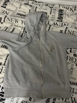 Grå Moncler hoodie med dragkedja - Snygg grå hoodie från Moncler med dragkedja framtill och två stora fickor. Hoodien har en klassisk Moncler-logga på bröstet och en mjuk, bekväm känsla. Perfekt för en avslappnad streetwear-look.