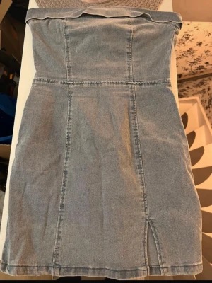 Axelbandslös jeansklänning - Trendig axelbandslös jeansklänning i ljusblå denim med slits framtill och dragkedja bak. Klänningen har en figurnära passform och kort längd, endast använd en gång :)