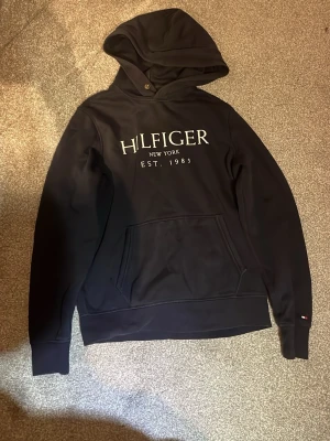 Svart Hilfiger hoodie med tryck - Svart hoodie från Tommy Hilfiger med stor vit logga och texten 'HILFIGER NEW YORK EST. 1985' på bröstet. Klassisk känguruficka framtill och liten Hilfiger-flagga på ärmen. Mjuk bomullsblandning och huva med dragsko. Perfekt för en avslappnad streetwear-look.