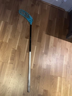 Unihoc innebandyklubba Ultralight - Säljer en Unihoc innebandyklubba Ultralight med svart skaft, vit grepptejp och ett blått blad. Klubban är lätt och smidig, perfekt för snabba spelare som vill ha bra bollkontroll. Bladet har ett modernt mönster och klubban har stilrena detaljer.