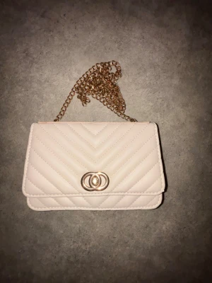 Ljusbeige quiltad kuvertväska - Snygg ljusbeige kuvertväska med quiltat mönster och guldfärgad kedja. Väskan har ett elegant dubbelring-spänne framtill och ett innerfack med dragkedja. Perfekt att bära crossbody eller i handen för en trendig look.