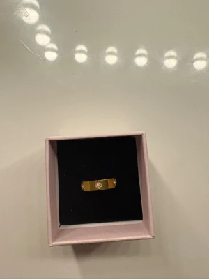 Guldfärgad ring med stenar - Snygg guldfärgad ring med en rak, slät design och tre gnistrande stenar infällda framtill. Ringen levereras i en rosa ask och har en modern, minimalistisk look som passar perfekt till din stil.