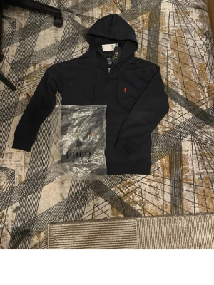 Svart hoodie från Polo Ralph Lauren - Snygg Svart hoodie från Polo Ralph Lauren med klassisk huva och dragsko. Ikoniska röda loggan broderad på bröstet. Tillverkad i mjukt bomullsmaterial och har ribbade muddar vid ärmslut och nederkant. Perfekt för en avslappnad och stilren look.