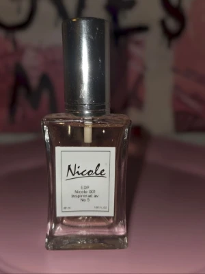Nicole 001 Eau de Parfum 30ml - Nicole 001 Eau de Parfum är en parfym inspirerad av No 5. Flaskan är rektangulär i genomskinligt glas med en silverfärgad kork och vit etikett. Doften är elegant och klassisk, perfekt för dig som gillar ikoniska parfymer.
